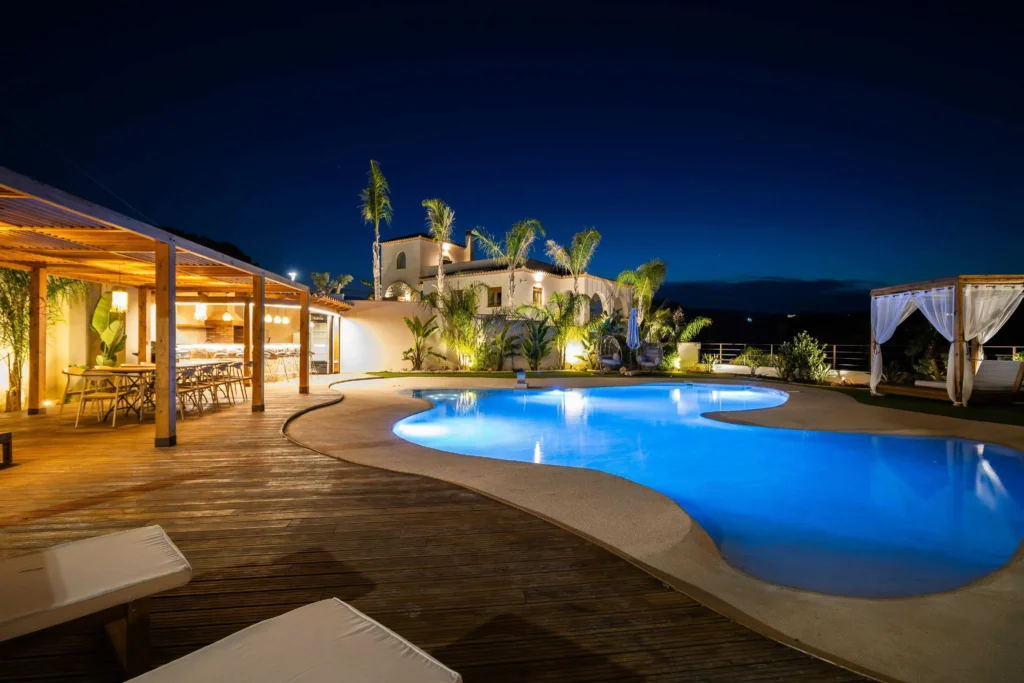 Stunning tropical holiday villa featuring a curved private pool with underwater LED lights, wooden pergola with LED accent lighting, and palm trees. Energy-saving LED lighting creates a romantic, relaxing atmosphere for evening relaxation.