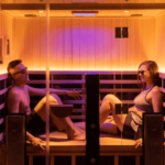 Two adults sit across from each other in a glass-enclosed sauna, wearing swimsuits and relaxing in warm light.