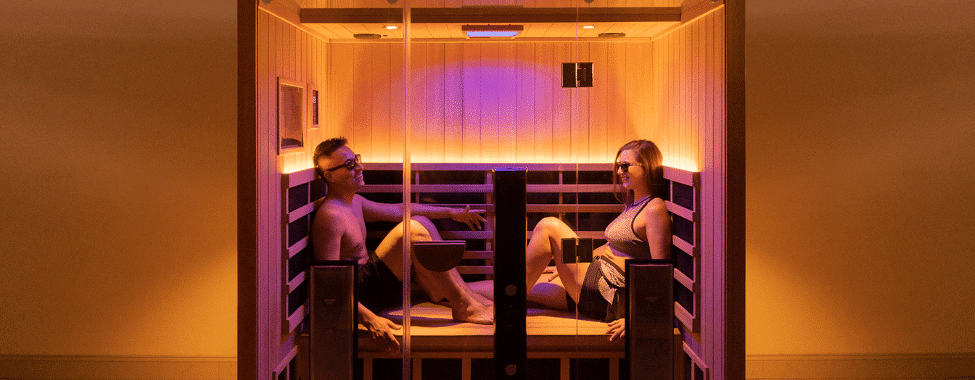 Two adults sit across from each other in a glass-enclosed sauna, wearing swimsuits and relaxing in warm light.