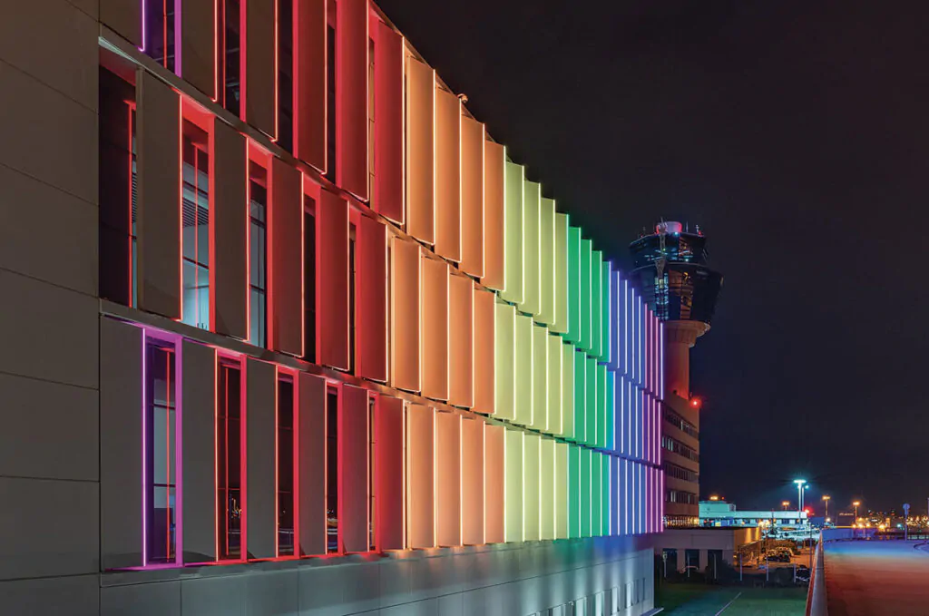 Large-scale airport terminal building facade illuminated at night with colorful RGB LED strip lights creating a gradient rainbow effect from purple to red, orange, yellow, green and blue, with air traffic control tower visible in background
