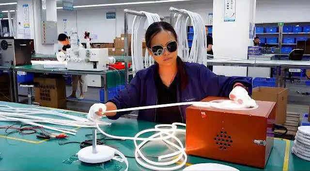 Female technician in a blue uniform and sunglasses assembles white tubing at a green factory workstation, with a brown electrical box and coiled cables nearby in a manufacturing setting.