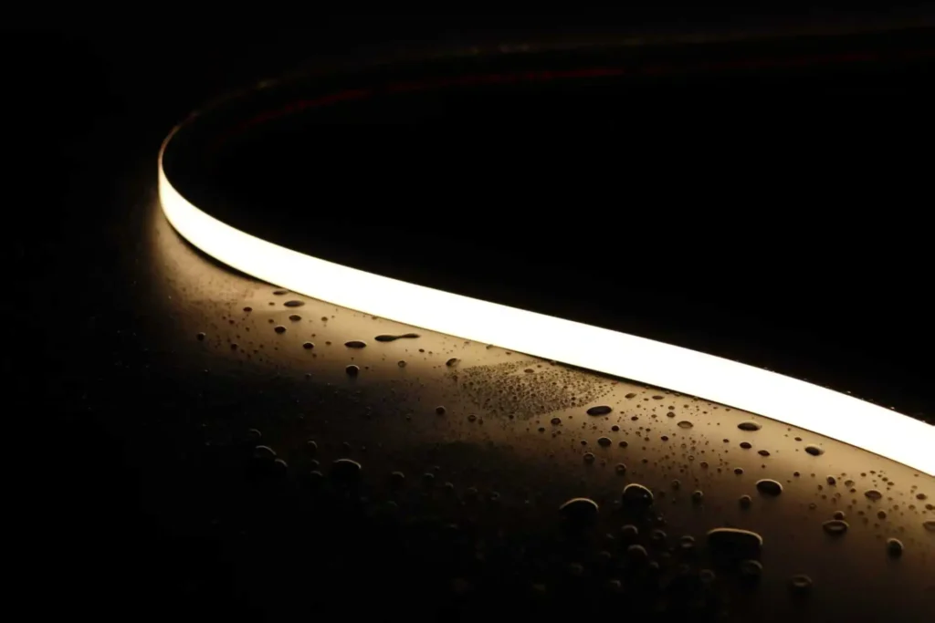 Close-up of a warm white waterproof silicone LED strip light glowing brightly with water droplets scattered across its surface on a dark background, demonstrating IP67 water resistance and the strip's ability to maintain illumination in wet conditions