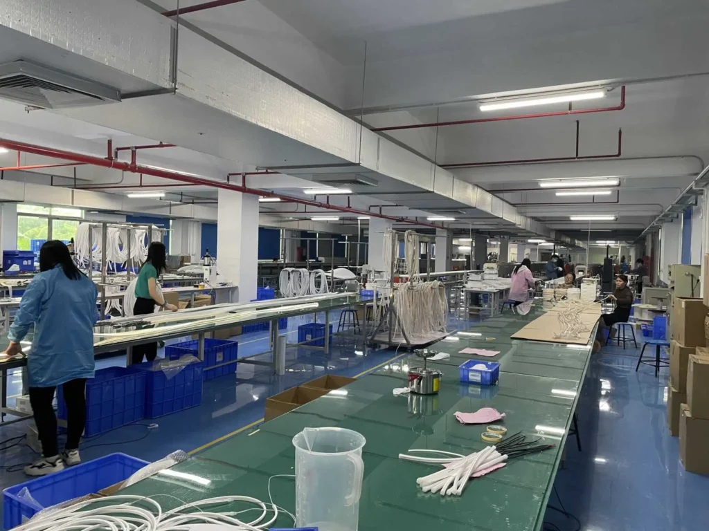 oem-led-neon-flex-manufacturing-workshop » Custom LED Strip Lights Manufacturer | Silicone Neon Flex Supplier China » Custom LED Strip Lights Manufacturer | Silicone Neon Flex Supplier China Industrial sewing/assembly workshop with workers at long workbenches and blue plastic bins nearby, overhead piping visible.