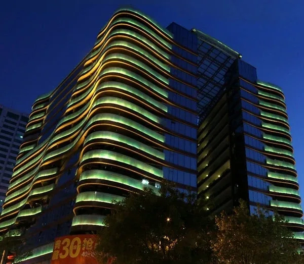 Night view of a high-rise building with wavy illuminated green and yellow horizontal bands along the facade, at dusk/night.